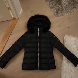 Svart dunjacka från Chiara Forthi nyskick  - Snygg svart dunjacka från Chiara Forthi med pälskantad huva och dragkedja. Jackan har en quiltad design och en praktisk ficka med dragkedja på ärmen. Perfekt för kyliga dagar!