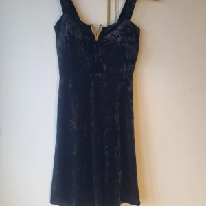 Svart sammet klänning - Elegant svart klänning i mjuk sammet med en stilren V-detalj vid halsringningen. Vadderade kupor på klänningen. Klänningen har en figurnära passform och är ärmlös, vilket gör den perfekt för festliga tillfällen.