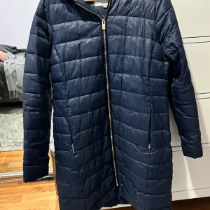 Mörkblå quiltad jacka med huva - Snygg mörkblå quiltad jacka med dragkedja och huva. Perfekt för kyliga dagar. Jackan har långa ärmar och praktiska fickor med dragkedja framtill. Använd några gånger, lång