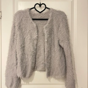 Grå fluffig kofta - Supermysig grå kofta i fluffigt material, perfekt för kyliga dagar. Den har en öppen design utan knappar och långa ärmar. Perfekt för att hålla sig varm och stilren samtidigt! 