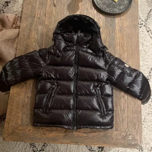 Svart dunjacka från Moncler - Snygg svart dunjacka från Moncler med glansig finish och huva. Jackan har en dragkedja framtill och praktiska fickor med knappar. Perfekt för kyliga dagar.