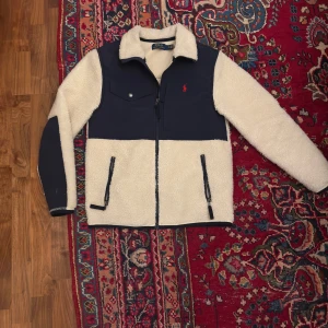  Ralph Lauren fleece jacka  - Säljer en snygg fleece jacka från Polo Ralph Lauren i beige och marinblått. Jackan har dragkedja framtill, två fickor med dragkedja och en bröstficka. Perfekt för kyliga dagar med sin mjuka och varma design. Dom är även storlek XS men kan passa S perfekt 