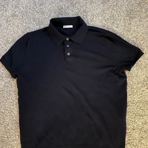 Svart pikétröja från Zara - Säljer en stilren svart pikétröja från Zara. Tröjan har korta ärmar och en klassisk krage med tre knappar. Perfekt för en avslappnad men ändå snygg look.