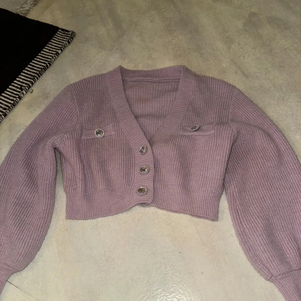 Säljer en söt lila stickad kofta med glittriga knappar framtill. Koftan är croppad och har långa ärmar med ribbade muddar. Perfekt för att lägga till lite glitter i vardagen!. Neuletakit & Villapaidat.