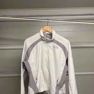 Vit och grå vindjacka från The North Face - Säljer en snygg och praktisk vit och grå vindjacka från The North Face. Jackan har en dragkedja framtill och är långärmad. Perfekt för vår och höst när det blåser lite extra. Den är i bra skick och redo för nya äventyr! Säljes i befintligt skick. Storlek S/M. 