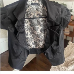 Svart bolero från Vila - Snygg svart bolero från Vila med ett elegant blommigt mönster på insidan. Perfekt för både vardag och fest. Kavajen har en klassisk design med knappar framtill och en stilren passform.