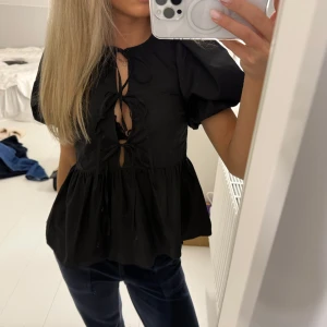 Svart blus med puffärmar - Snygg svart blus med puffiga kortärmar och trendig snörning framtill. Perfekt för både vardag och fest! Den har en luftig passform och är superbekväm att bära. Passar bra till jeans eller kjol för en chic look.