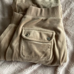 Beige mjukisbyxor med fickdetaljer - Säljer ett par bekväma beige mjukisbyxor med elastisk midja och snörning. Byxorna har praktiska fickor med knappdetaljer och en broderad logotyp. Perfekta för en avslappnad stil.