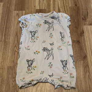 Beige babyklänning med Disney-motiv från H&M - Söt beige babyklänning från H&M med Disney-motiv av Bambi och blommor. Klänningen är kortärmad och tillverkad i ekologisk bomull, perfekt för små barn. Den har praktiska knappar framtill för enkel påklädning.