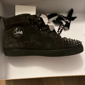 Svarta sneakers med nitar från Christian Louboutin - Snygga svarta sneakers i mocka från Christian Louboutin med ikoniska röda sulor. Skorna har snörning och är dekorerade med nitar på tån för en edgy look. Perfekta för att ge din outfit en unik touch.