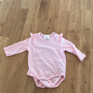 Rosa långärmad body från Bonprix - Söt rosa långärmad body från Bonprix med volangdetaljer vid axlarna. Tillverkad i ekologisk bomull, perfekt för de minsta. Praktiska knappar vid axeln och grenen för enkel påklädning.