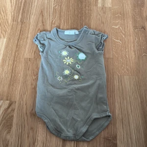 Grå body med blommönster - Söt grå body från Åhlens med korta ärmar och tryck av blommor i vitt och gult på framsidan. Praktiska knappar vid axeln och i grenen för enkel på- och avklädning. Perfekt för de små under varmare dagar.