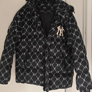Svart pufferjacka från MLB - XL size  - Snygg svart pufferjacka från MLB med ett mönster av vita 'NY' logotyper. Jackan har en dragkedja och knappar framtill samt en huva för extra värme. Perfekt för kyliga dagar.