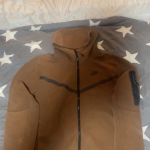 Brun Nike tech fleece  - Säljer en brun jacka från Nike med dragkedja och huva. Jackan har en stilren design med svarta detaljer och Nike-logga på bröstet. Perfekt för höst och vår.