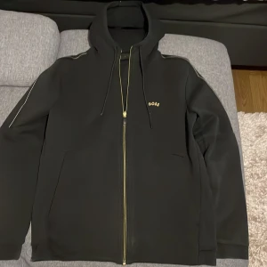 Svart hoodie från BOSS - Säljer en stilren svart hoodie från BOSS i mycket bra skick. Den har en dragkedja framtill och en liten BOSS-logga på bröstet. Perfekt för höst och vår, med långa ärmar och en bekväm passform. Grym för både chill och casual outfits!