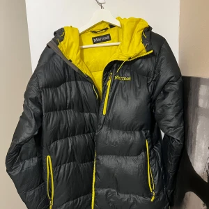 Varm svart och gul dunjacka från Marmot - Säljer en snygg svart dunjacka från Marmot med en cool gul insida och detaljer. Jackan har en praktisk huva och dragkedja framtill. Perfekt för kalla vinterdagar och höstpromenader. Den är i mycket bra skick.