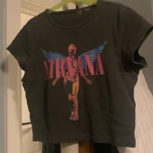 Svart Nirvana t-shirt - Säljer en cool mörkgrön/svart Nirvana t-shirt med färgglatt tryck av en ängel och bandets namn i rosa. T-shirten är croppad och kortärmad, perfekt för en avslappnad stil. Passar bra till jeans eller kjol för en rockig look. Hyfsat tight. 