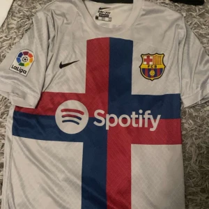 Vit FC Barcelona fotbollströja - Säljer en vit FC Barcelona fotbollströja med korta ärmar. Den har ett stort kors i rött och blått på framsidan och La Liga-märke på ärmen. På baksidan står det 'Lewandowski' och nummer 9. Perfekt för fans av laget och spelaren!