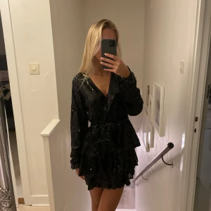 Svart glittrig klänning - Snygg svart klänning med paljetter och volangdetaljer. Den har en v-ringad design och långa ärmar, perfekt för en festlig kväll.