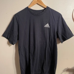 Svart t-shirt från Adidas - Säljer en klassisk svart t-shirt från Adidas i storlek M. Den är i bra skick och har en vit Adidas-logga på bröstet. Perfekt för träning eller vardagsbruk. Materialet känns mjukt och bekvämt. Passar till det mesta och är ett måste i garderoben!
