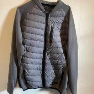 Grå jacka från MyProtein - Säljer en grå jacka från MyProtein i storlek L. Den har en quiltad design med dragkedja framtill och praktiska fickor. Perfekt för höst och vår, med långärmade ärmar och en sportig look. Jackan är i bra skick och redo för nya äventyr!