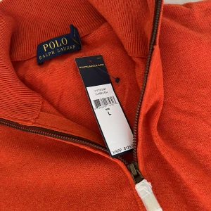 Oanvänd tröja från Polo Ralph Lauren i merinoull  - Säljer en snygg orange tröja från Polo Ralph Lauren. Den har en dragkedja vid halsen och det klassiska Polo-logotypen broderad på bröstet. Perfekt för höst och vår med sin mjuka och bekväma passform. Tröjan är i oanvänd och redo för nya äventyr! 100% merinoull. Ett mikroskopiskt hål nedtill i mudden. Jag har en likadan fast blå som jag upplever lite stor i storleken.