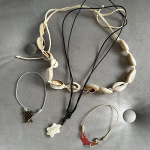 Set med strandinspirerade smycken - Säljer ett set med fyra strandinspirerade smycken. Det finns två halsband och två armband. Ett halsband är gjort av vita snäckor med en justerbar snodd, perfekt för sommarens strandhäng. Det andra halsbandet har en svart snodd med ett vitt hänge i form av en sköldpadda. Ett av armbanden har en silverfärgad snodd med ett sköldpaddshänge, medan det andra har en guldig snodd med ett rött delfinhänge och en liten silverpärla. Alla smycken är i bra skick och redo för nya äventyr!