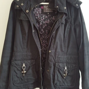 Svart jacka med blommigt foder - Säljer en snygg svart jacka från Esprit med ett blommigt foder i rosa och vitt. Jackan har en huva och praktiska fickor med spännen framtill. Perfekt för höst och vår!