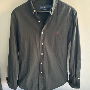 Svart skjorta från Ralph Lauren - Säljer en snygg svart skjorta från Ralph Lauren i slim fit. Skjortan har långa ärmar och klassiska knappar framtill. Den är gjord i bomull och har en röd broderad logga på bröstet. Perfekt för både vardag och fest!