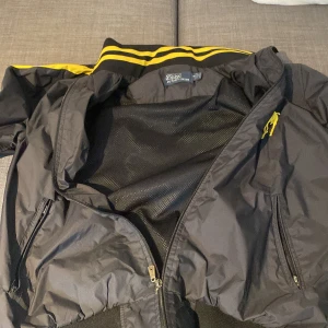 Svart vindjacka från Polo Ralph Lauren - Säljer en snygg svart vindjacka från Polo Ralph Lauren. Jackan har gula detaljer längs ärmarna och en stor gul logga på bröstet. Den är långärmad med dragkedja framtill och har ribbade muddar vid ärmslut och nederkant. Perfekt för vår och höst! skaftet på dragkedjan på bild tre är av men den går att stänga och fungerar felfritt!