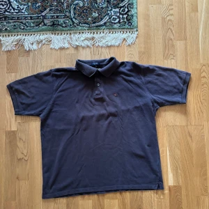 Mörkblå pikétröja från Burberry - Säljer en snygg mörkbrun pikétröja från Burberry i storlek XL. Tröjan är i bomull och har korta ärmar med klassisk krage och knappar. Den har en liten broderad logga på bröstet och den ikoniska Burberry-rutiga detaljen på insidan av kragen.