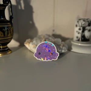 Lila holografisk klistermärke - Ett supergulligt lila klistermärke med en holografisk effekt som ger det en cool glans. Perfekt för att dekorera din laptop, anteckningsbok eller vad du vill! Det har en söt liten ansikte som gör det extra charmigt.