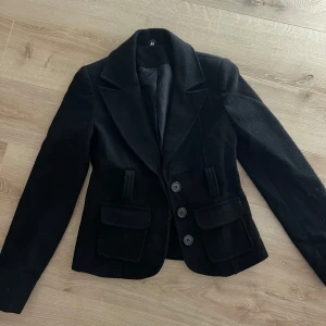 Svart kavaj med knappar - Snygg svart kavaj med tre knappar framtill och klassisk krage. Den har två fickor med lock och är perfekt för en stilren look. Passar bra till både vardag och mer formella tillfällen.