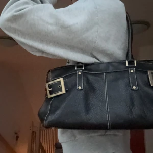Svart axelväska med spännen - Snygg svart axelväska med stiliga spännen och dragkedja. Perfekt för att bära med sig det viktigaste med stil. Väskan har en klassisk design som passar till många olika outfits.