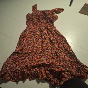 Blommig klänning med volangdetaljer - Charmig blommig klänning med volangdetaljer och oneshoulder-design. Perfekt för sommarens varma dagar med sitt färgglada mönster och luftiga passform.