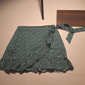 Grön blommig omlottkjol - Charmig grön omlottkjol med vita blommor och volangdetaljer. Kjolen har ett knytband i midjan för justerbar passform och en lekfull volangkant som ger extra stil. Perfekt för en somrig look!