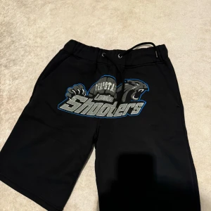Svarta shorts med tryck - Coola svarta shorts med ett stort tryck av en panter och texten 'Shooters'. De har en bekväm passform med snörning i midjan. Perfekta för en avslappnad stil.