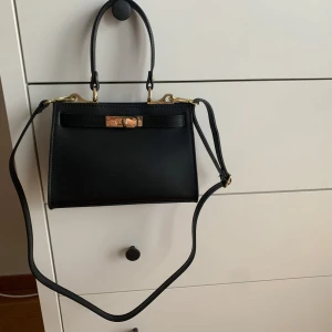 Svart handväska med gulddetaljer - Skinnväska ”Latina” från Latalia. Knappt använd så i mycket fint skick. Svart insida med liten innerficka. Reglerbart axelband. Väskan är 23 x 7 x 15 cm. Finns på fler sidor.