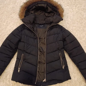 Svart vinterjacka från Everest - Säljer en svart dunjacka från Everest med avtagbar fuskpäls på huvan. Jackan har dragkedja framtill och två fickor med dragkedjor. Perfekt för alla olika snöiga och kalla dagar med sin quiltade design och långa ärmar.