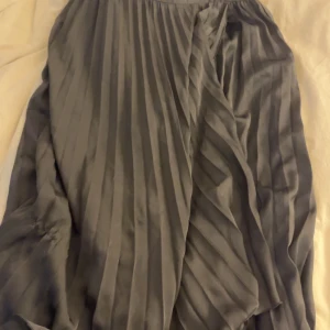 Grå omlottkjol från Gina tricot  - Snygg grå omlottkjol från Gina tricot i ett elegant plisserat tyg. Kjolen har en omlottdesign som ger en fin rörelse och stil. Perfekt för att skapa en chic look.