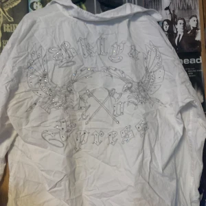 Vit skjorta med tryck och stenar - Snygg vit skjorta med unikt tryck och glittrande stenar på både fram- och baksida. Skjortan har långa ärmar och knappar framtill. Perfekt för att sticka ut med en cool och edgy stil.