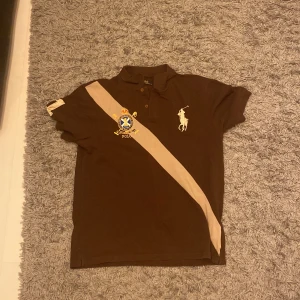 Brun pikétröja från Ralph Lauren - Säljer en snygg brun pikétröja från Ralph Lauren med en beige diagonal rand och broderad logga. Tröjan har korta ärmar och klassisk krage. Perfekt för en stilren look.den säger xxl men passar som en L