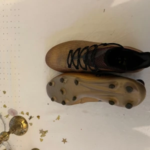 Guldiga fotbollsskor från Adidas - Snygga guldiga fotbollsskor från Adidas med svarta detaljer och snörning. Skorna har dobbar för bra grepp på planen och en stilren design med Adidas-loggan på sidan. Perfekta för fotbollsspelare som vill sticka ut på planen.