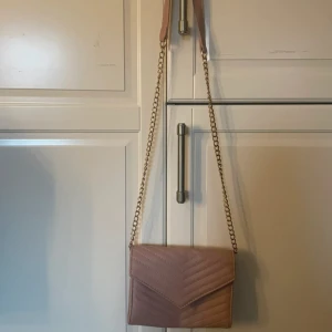 Beige axelväska med kedja - Snygg beige axelväska med kedjedetaljer och quiltad design. Väskan har ett lock med magnetknäppning och en justerbar axelrem. Perfekt för att bära dina nödvändigheter med stil.