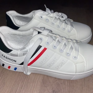 Vita Fashion Sneaker - Snygga vita sneakers med texten 'FASHION' och 'SPORT' på sidan. Skorna har snörning och färgglada detaljer i rött, blått och svart. Perfekta för en sportig look.