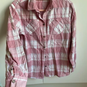Rutig rosa och vit skjorta - Säljer en snygg rutig skjorta i rosa och vitt med långa ärmar och knappar framtill. Perfekt för en avslappnad stil. Skjortan har två bröstfickor och en klassisk krage.