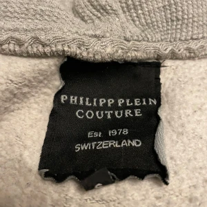 Grå hoodie från Philipp Plein - Säljer en grå hoodie från Philipp Plein med en cool dödskalletryck på ryggen. Tröjan har dragkedja framtill och en bekväm huva. Perfekt för en avslappnad stil.