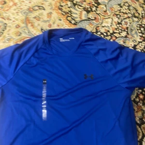 Blå t-shirt från Under Armour - Säljer en blå t-shirt från Under Armour med korta ärmar och rund hals. Perfekt för träning eller en aktiv dag. Den har en liten logotyp på bröstet och är tillverkad i ett lätt och andningsbart material.