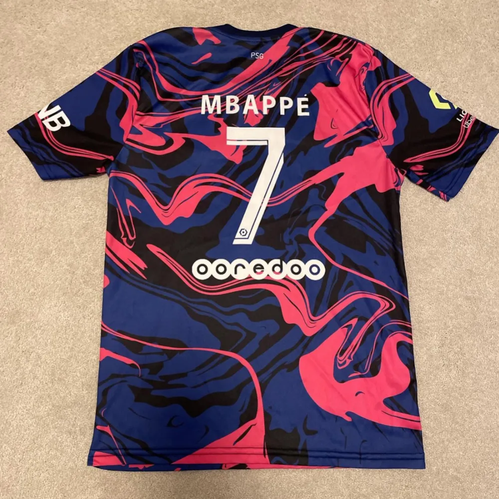 Snygg PSG-tröja i samarbete med Jordan. Den har ett unikt mönster i blått, rosa och svart med vit text.. T-paidat.