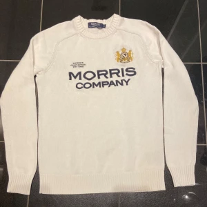 Vit tröja från Morris - Säljer en stilren vit tröja från Morris med broderad logotyp och text på framsidan. Tröjan är tillverkad i 100% bomull och har långa ärmar med ribbade muddar. Perfekt för en klassisk look.